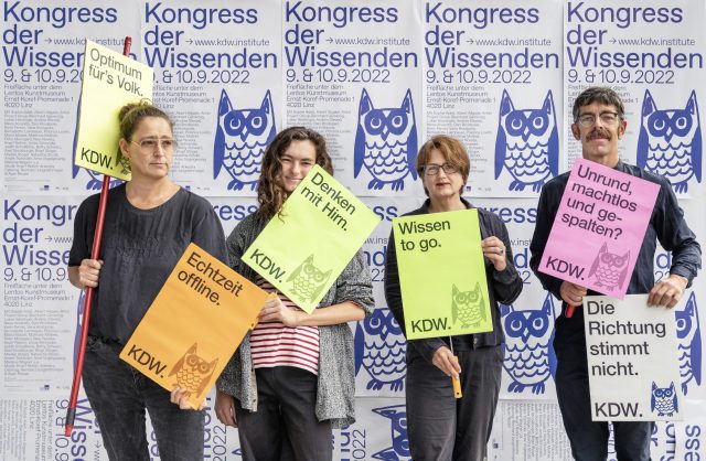 Eine Reihe von Menschen steht mit Plakaten in der Hand vor einer Wand mit Flyern des Kongress der Wissenden