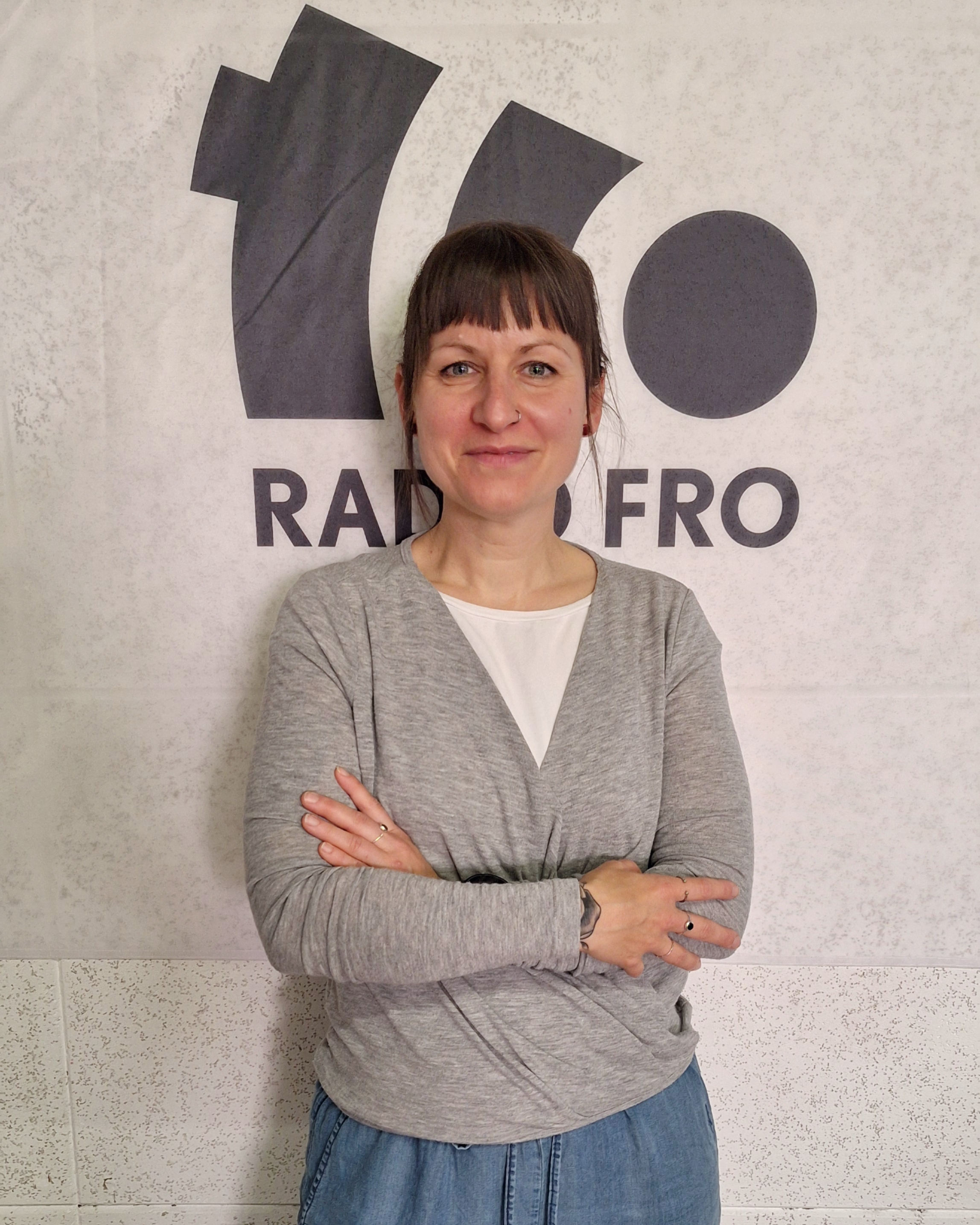 Eine Frau steht mit verschränkten Armen vor einer weißen Wand, auf der in schwarz das Radio FRO Logo abgebildet ist. Sie lächelt in die Kamera.