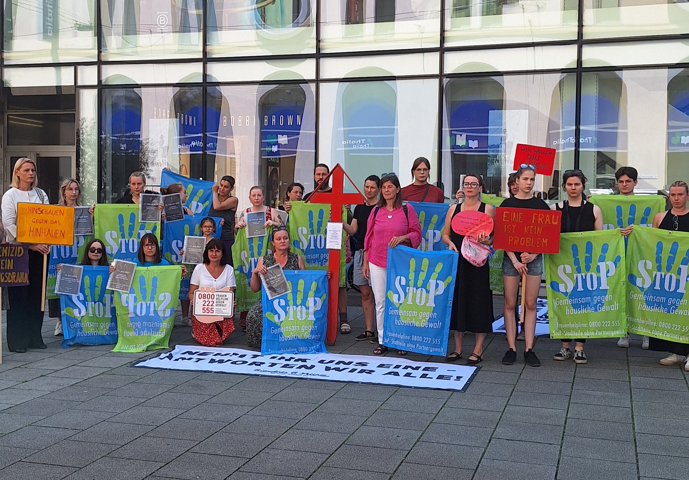 Eine Gruppe an Frauen hält grün-blaue Banner mit dem Stadtteile ohne Partnergewalt in die Höhe. Außerdem halten ein paar Personen Schilder hoch, auf denen Femizide angeprangert werden. 