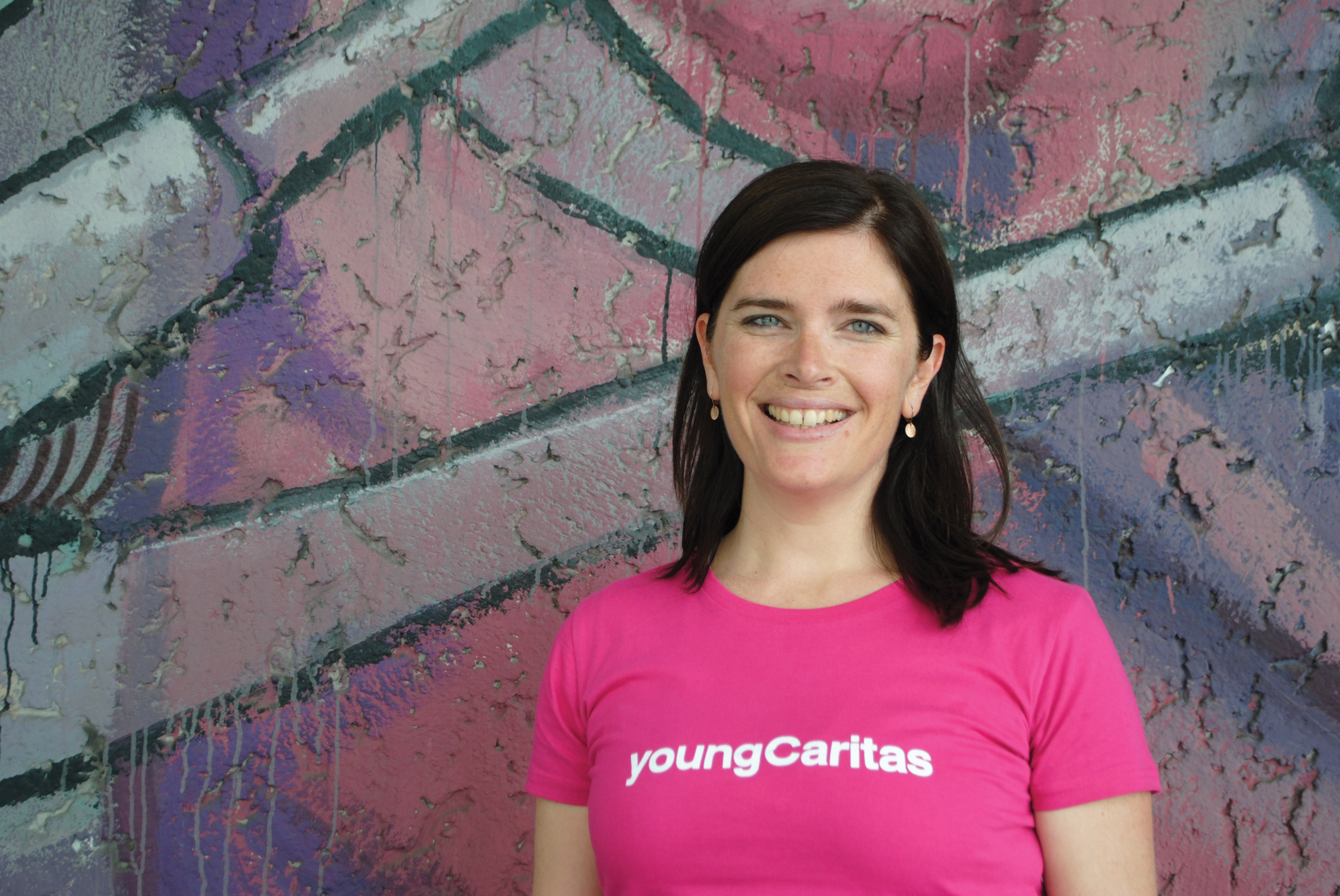 Eine Frau steht vor einer Wand, auf der Ausschnitte eines Graffiti zu sehen sind. Sie lächelt in die Kamera und auf ihrem pinken Shirt steht in weißer Schrift: Young Caritas.