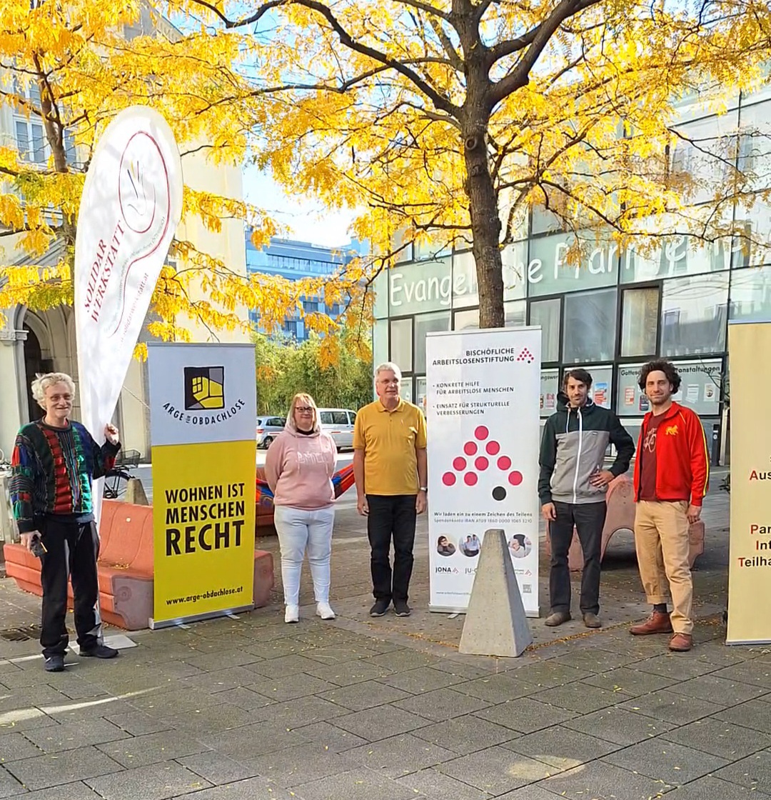 Personen stehen am Martin Luther Platz in Linz und haben sich neben Roll-Ups von verschiedenen zivilgesellschaftlichen Organisationen aufgestellt.