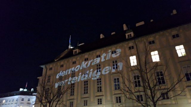 Auf eine Gebäudefront am Ballhausplatz in Wien wird in weißer Schrift meterhoch hinprojiziert: Demokratie verteidigen